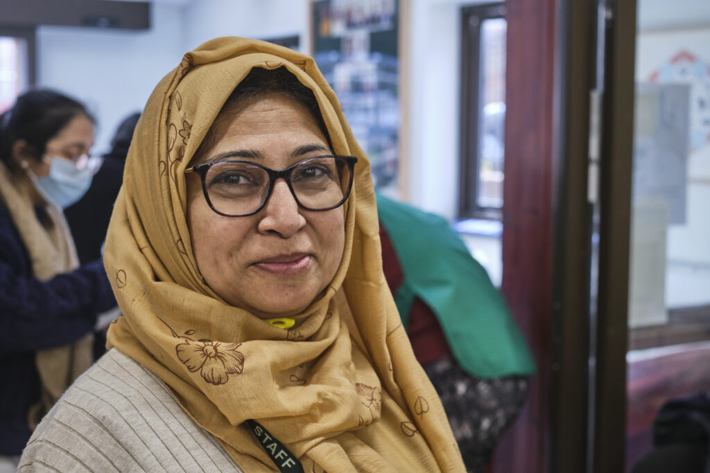 A Muslim woman, wearing a yellow hijab, black glasses and a brown top, is smiling at the camera