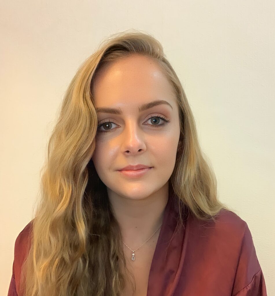 A photo of gabrielle with long, blonde hair, wearing a dark red top. She is sat in front of a white wall.