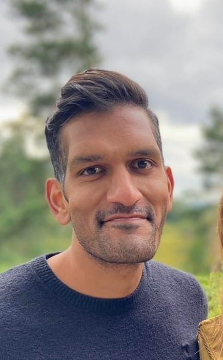 A photo of Ketan with short, brown hair, wearing a grey jumper. He is stood in front of some trees.