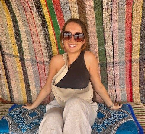 A photo of rachel wearing cream trousers and a black top, sat in front of a striped canvas background. She is wearing sunglasses and smiling