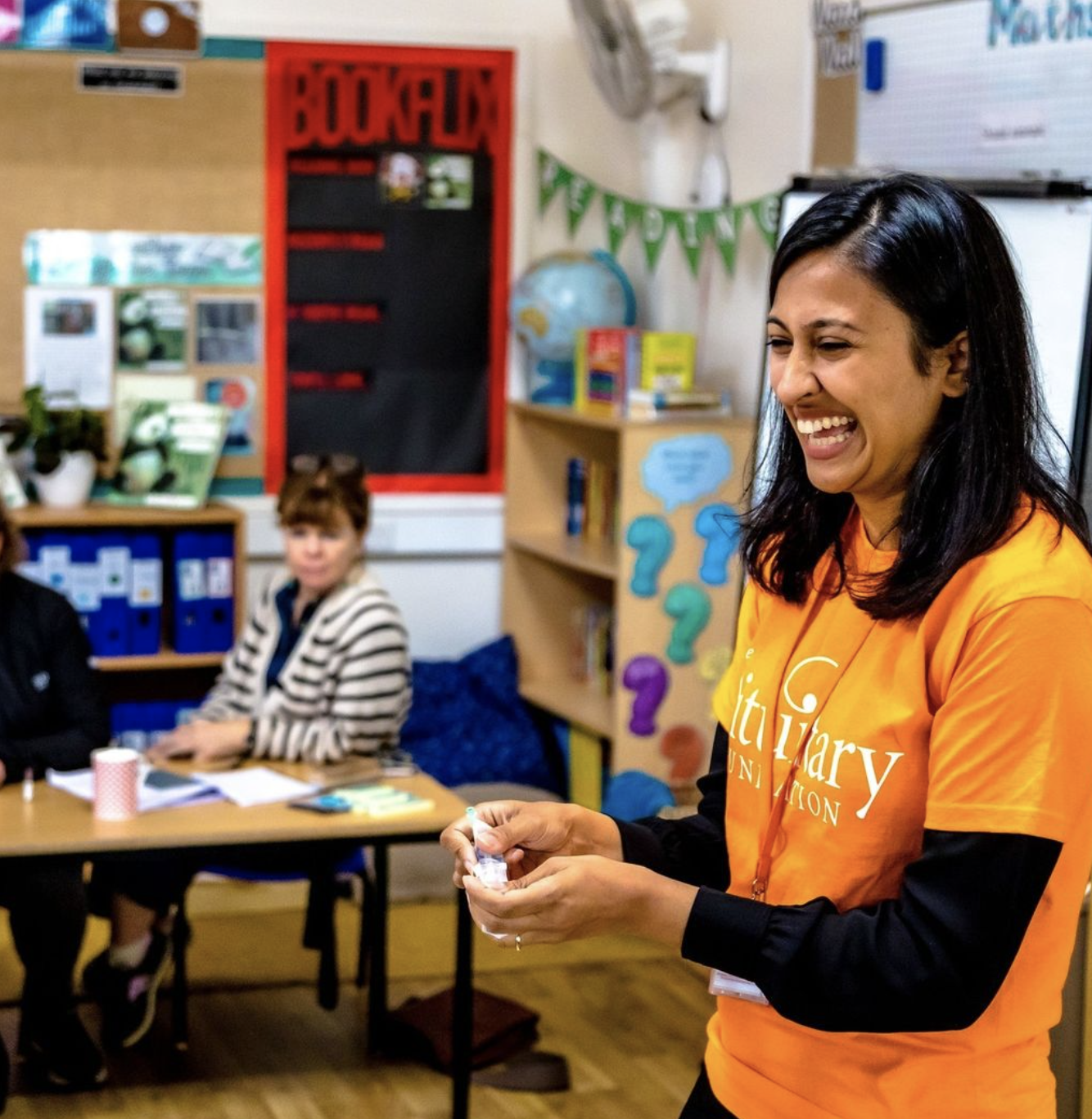 Darshna is wearing an orange pituitary foundation top and is stood in front of a classroom, leading an educational session