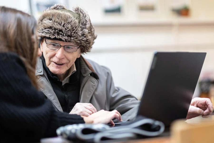 An older man is sat behind a latptop, talking to younger woman.